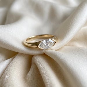 Puede incluir: Un anillo de oro con una sola piedra preciosa transparente de talla marquesa engastada horizontalmente en una banda lisa y pulida. El engaste sujeta la piedra de forma segura entre dos puntas metálicas cónicas y definidas.