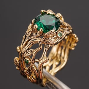Puede incluir: Un anillo de oro con una gran piedra preciosa verde de corte redondo en el centro, rodeado por una banda diseñada para parecer ramas y hojas entrelazadas, con pequeñas piedras verdes incrustadas en la estructura metálica.