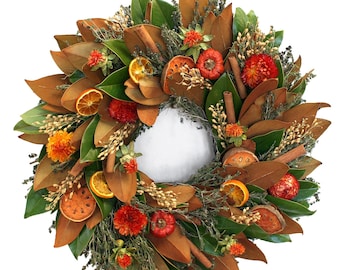 Könnte beinhalten: Ein kreisförmiger Kranz aus natürlichen Elementen. Er besteht aus großen, braunen Magnolienblättern, grünen Blättern, Orangenscheiben, Zimtstangen und kleinen orangefarbenen und roten Blüten. Der Kranz hat eine weiße Mitte.