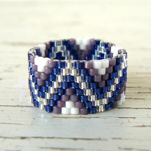 May include: A beaded ring with a geometric pattern. The ring features a repeating pattern of blue, silver, white, and purple beads. The ring is approximately 2 cm wide.