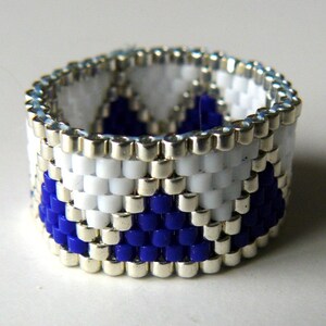 May include: A beaded ring with a geometric pattern in white and blue beads. The ring is made with silver-colored beads.