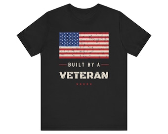 Op de afbeelding: Een zwart t-shirt met korte mouwen voorzien van een afbeelding van de verweerde Amerikaanse vlag boven de tekst BUILT BY A VETERAN in witte hoofdletters, met vijf kleine rode sterren gecentreerd onder de tekst op de voorzijde.