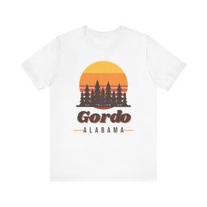Puede incluir: Una camiseta blanca de manga corta con un gráfico de un atardecer con rayas horizontales naranjas y amarillas detrás de una silueta marrón oscuro de pinos. Debajo de los árboles, el texto dice Gordo y ALABAMA.