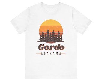 Op de afbeelding: Een wit t-shirt met korte mouwen voorzien van een afbeelding van een zonsondergang met horizontale oranje en gele strepen achter een donkerbruin silhouet van dennenbomen. Onder de bomen staat de tekst Gordo en ALABAMA.