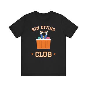 Puede incluir: Una camiseta negra de manga corta con un gráfico de una persona sumergiéndose en un contenedor naranja con las piernas hacia afuera. El texto dice BIN DIVING CLUB en letras naranja claro con dos pequeñas estrellas.