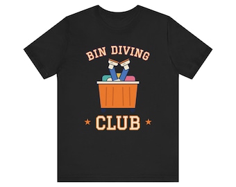 Op de afbeelding: Een zwart t-shirt met korte mouwen en een afbeelding van een persoon die in een oranje bak duikt met de benen naar buiten. De tekst BIN DIVING CLUB staat in lichtoranje letters met twee kleine sterren ernaast.