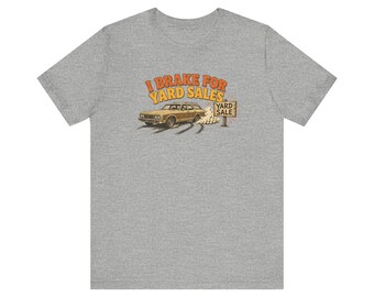 Op de afbeelding: Een grijs gemêleerd T-shirt met korte mouwen en een afbeelding van een beige stationwagen die remt voor een houten bord met de tekst YARD SALE. Boven de auto staat de tekst I BRAKE FOR YARD SALES in oranje en gele letters.