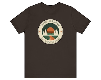Op de afbeelding: Een donkerbruin t-shirt met korte mouwen en een cirkelvormige afbeelding op de borst. Het ontwerp toont een zonsondergang boven een bos en een kronkelende weg, met de tekst "WEST ALABAMA" en "EST. COUNTRY ROADS" op een crèmekleurige rand.