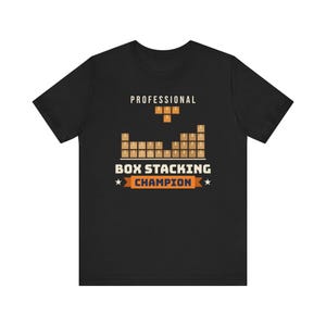 Puede incluir: Una camiseta negra de manga corta con un gráfico de cajas marrones apiladas. El texto dice PROFESSIONAL BOX STACKING CHAMPION en letras blancas y naranjas, con una pequeña forma de bloque naranja sobre las cajas.