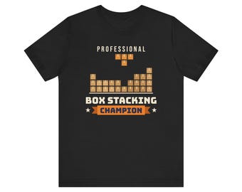 Op de afbeelding: Een zwart t-shirt met korte mouwen en een afbeelding van gestapelde bruine dozen. De tekst luidt PROFESSIONAL BOX STACKING CHAMPION in witte en oranje letters, met een kleine oranje blokvorm boven de gestapelde dozen.
