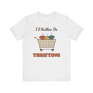 Puede incluir: Una camiseta gris claro de manga corta con un gráfico de un carrito de la compra lleno de tres camisas en perchas. Sobre el carrito está el texto I'd Rather Be y debajo la palabra THRIFTING en letras naranjas en negrita.