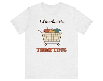 Op de afbeelding: Een lichtgrijs t-shirt met korte mouwen met een afbeelding van een winkelwagentje gevuld met drie shirts op kleerhangers. Boven het wagentje staat de tekst I'd Rather Be en onder het wagentje het woord THRIFTING in dikke oranje letters.