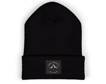 May include: A solid black knit beanie with a folded cuff. Centered on the cuff is a circular patch featuring a mountain range graphic and the text True North Alpine in white lettering against a dark background.