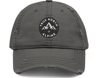 May include: A charcoal gray baseball cap with a distressed, worn-in finish. The front features a circular black patch with white embroidered text reading TRUE NORTH ALPINE, a white mountain peak graphic, and a small gold star icon.