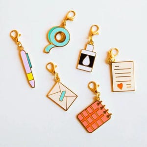 Pode incluir: Seis pingentes dourados com diferentes designs. Os pingentes incluem uma caneta rosa, um rolo de fita azul, uma garrafa branca com uma gota azul, um envelope branco com uma linha azul, um caderno rosa com um padrão de grade e uma nota branca com um coração vermelho.