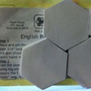 Op de afbeelding: Een close-up van zes grijze zeshoekige tegels, met instructies voor een Engels Paper Piecing-project. De instructies omvatten de stappen "Place and pin" en "Fold the unit".