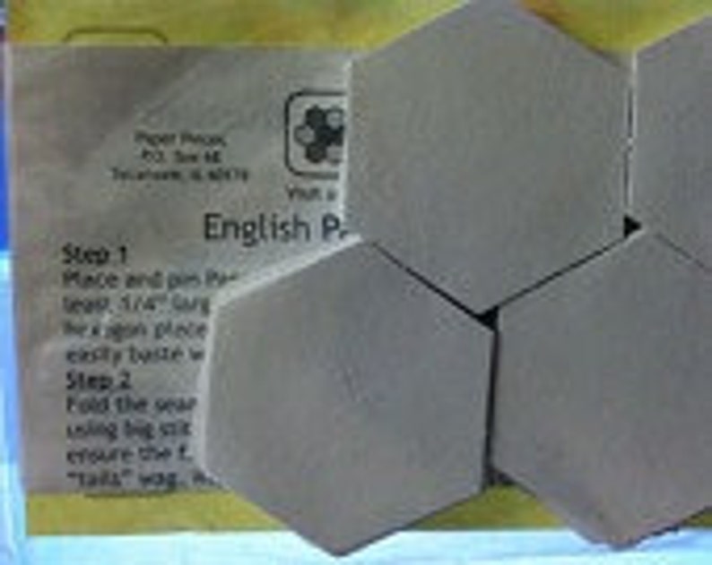 Op de afbeelding: Een close-up van zes grijze zeshoekige tegels, met instructies voor een Engels Paper Piecing-project. De instructies omvatten de stappen "Place and pin" en "Fold the unit".