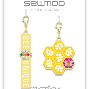 Puede incluir: Dos charms de cremallera dorados con temática de abejas. Un charm es una etiqueta rectangular con las palabras "Bee Unique" y el otro charm es un diseño de panal con una abeja en cada celda.