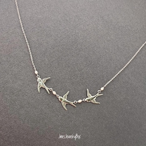 May include: A silver necklace with three swallow bird charms. The birds are linked by a fine chain with small, round beads. The necklace is shown against a grey backdrop.