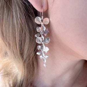 May include: Silver-toned floral drop earrings. Each earring features a series of stylized flower shapes, cascading down from the ear. The earrings have a delicate, elegant design, with a small bead at the bottom. The earrings are shown against a soft background.