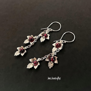 Può includere: Orecchini pendenti color argento con design a foglie, ciascuno con tre foglie e gemme rotonde rosso scuro. Gli orecchini pendono da un semplice gancio e sono esposti su uno sfondo scuro. Un gioiello.