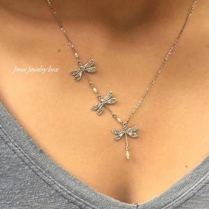 Puede incluir: Un collar de tono plateado con tres dijes de lib&eacute;lula dispuestos a lo largo de una cadena delicada. Cada lib&eacute;lula est&aacute; decorada con peque&ntilde;as piedras transparentes y diminutas perlas blancas. El texto Jmes Jewelry box es visible a la izquierda.