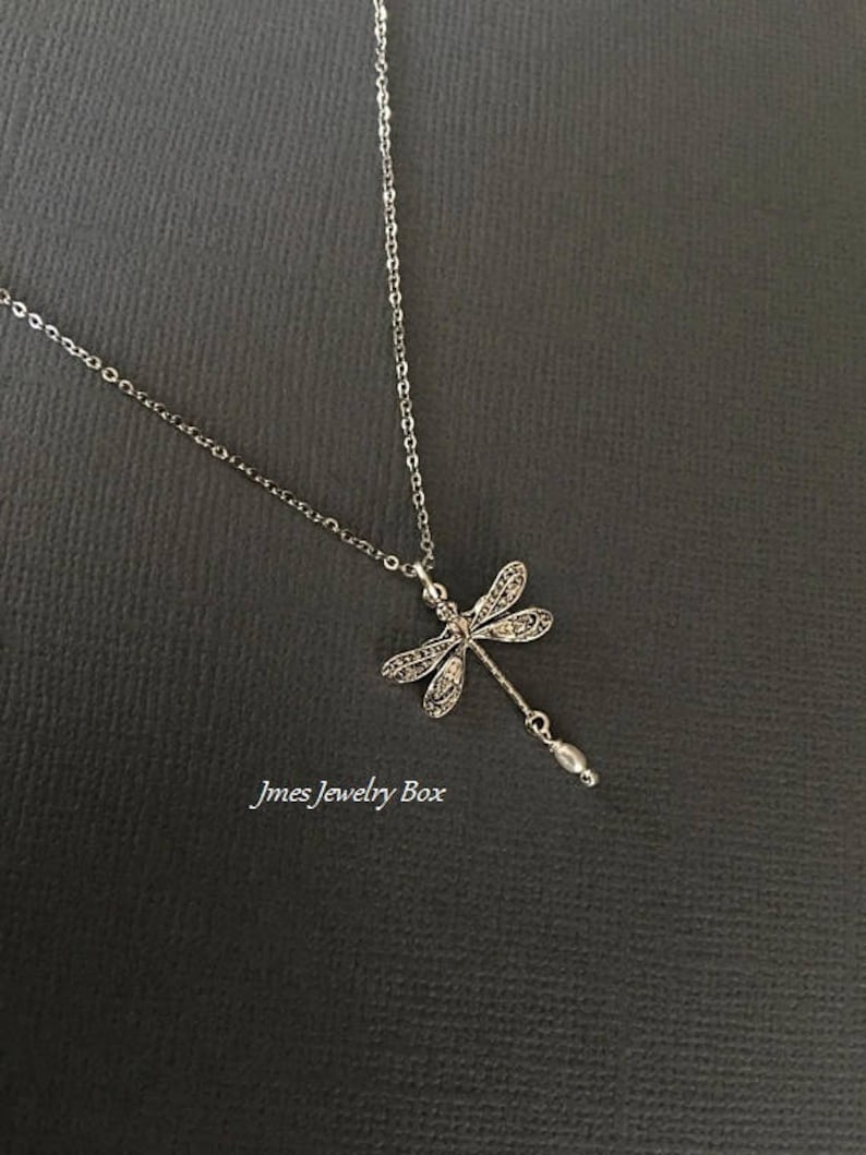 May include: A silver-toned necklace featuring a dragonfly pendant with intricate filigree wing details and a small dangling bead at the tail. The text Jmes Jewelry Box is visible in the lower left corner of the image. The chain is a simple link style.