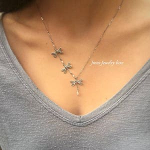 Puede incluir: Un collar de tono plateado con tres dijes de lib&eacute;lula dispuestos en l&iacute;nea diagonal, acentuados con peque&ntilde;as cuentas blancas. El texto Jmes Jewelry box es visible en el lado derecho de la cadena del collar sobre una camiseta gris con cuello en V.
