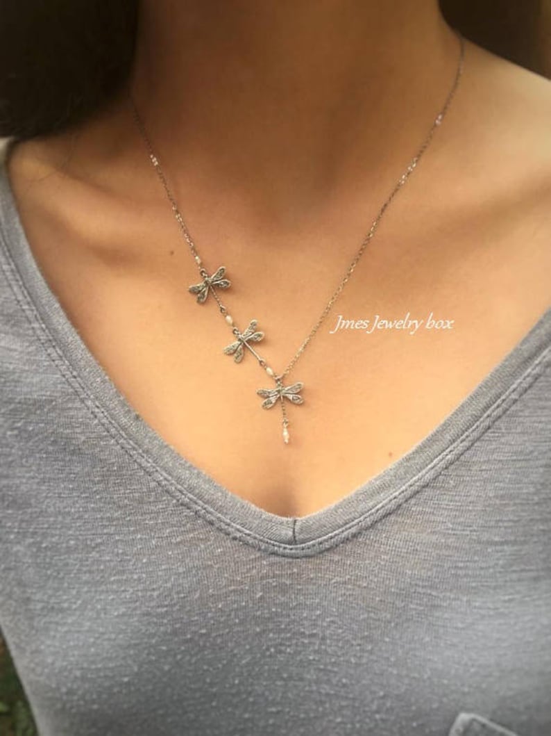 Puede incluir: Un collar de tono plateado con tres dijes de lib&eacute;lula dispuestos en l&iacute;nea diagonal, acentuados con peque&ntilde;as cuentas blancas. El texto Jmes Jewelry box es visible en el lado derecho de la cadena del collar sobre una camiseta gris con cuello en V.