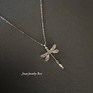 May include: A silver-toned necklace featuring a dragonfly pendant with intricate wing detailing and a small dangling bead at the tail. The text Jmes Jewelry Box is visible in the lower left corner of the image. The chain is a simple link style.