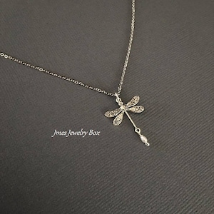 May include: A silver-toned necklace featuring a dragonfly pendant with intricate wing detailing and a small, dangling white pearl-like bead at the bottom. The text Jmes Jewelry Box is visible in the lower left area of the image.