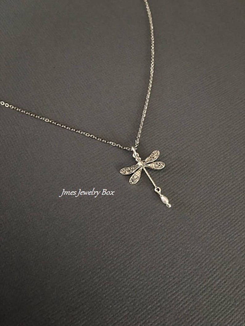 May include: A silver-toned necklace featuring a dragonfly pendant with intricate wing detailing and a small, dangling white pearl-like bead at the bottom. The text Jmes Jewelry Box is visible in the lower left area of the image.