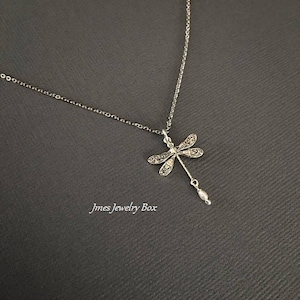 May include: A silver-toned necklace featuring a dragonfly pendant with intricate filigree-style wing detailing and a small, dangling white pearl-like bead at the base. The text Jmes Jewelry Box is visible in the lower left corner of the image.