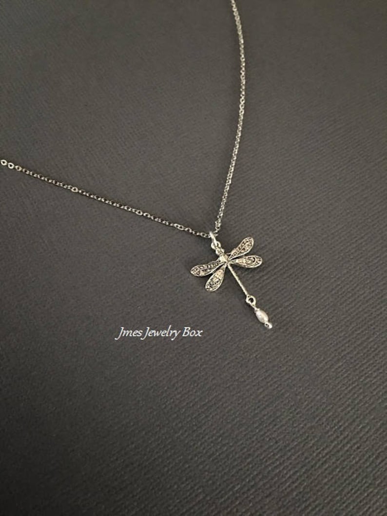 May include: A silver-toned necklace featuring a dragonfly pendant with intricate filigree-style wing detailing and a small, dangling white pearl-like bead at the base. The text Jmes Jewelry Box is visible in the lower left corner of the image.