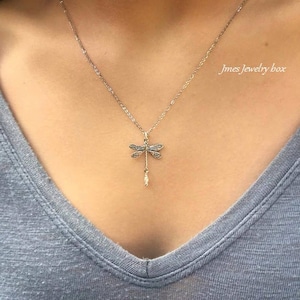 May include: A silver-toned necklace featuring a dragonfly pendant with intricate wing detailing and a small dangling pearl-like accent. The pendant hangs from a delicate chain. The text Jmes Jewelry box is visible in the upper right corner.