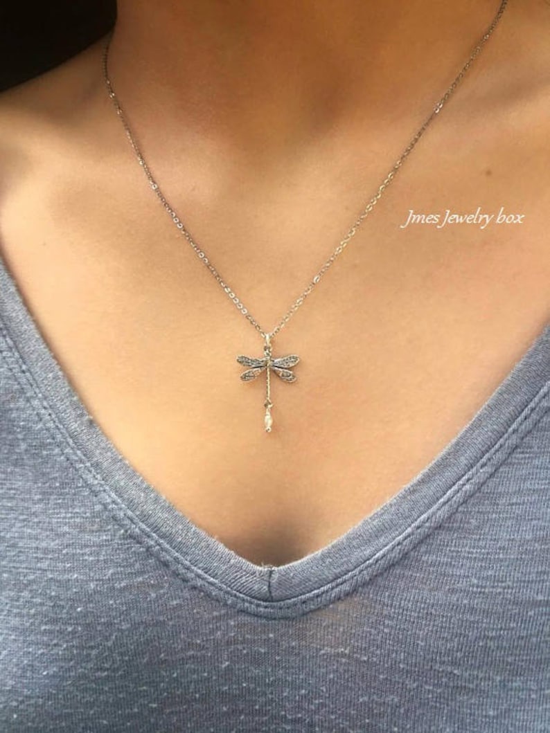 May include: A silver-toned necklace featuring a dragonfly pendant with intricate wing detailing and a small dangling pearl-like accent. The pendant hangs from a delicate chain. The text Jmes Jewelry box is visible in the upper right corner.