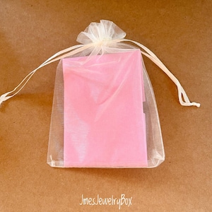 May include: A rectangular pink object is enclosed within a sheer, off-white organza drawstring pouch. The pouch features a gathered top with two thin matching ribbons tied in a bow. The text JmesJewelryBox is printed in white below the pouch.