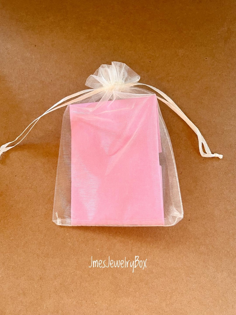May include: A rectangular pink object is enclosed within a sheer, off-white organza drawstring pouch. The pouch features a gathered top with two thin matching ribbons tied in a bow. The text JmesJewelryBox is printed in white below the pouch.
