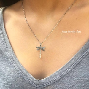 May include: A silver-toned necklace featuring a dragonfly pendant with intricate wing detailing and a small dangling white bead. The pendant hangs from a delicate chain. The text Jmes Jewelry box is visible in the upper right corner of the image.