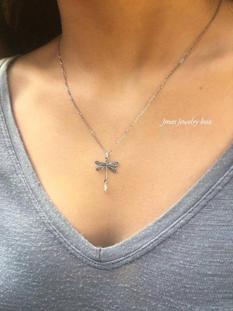 May include: A silver-toned necklace featuring a dragonfly pendant with intricate wing detailing and a small dangling white bead. The pendant hangs from a delicate chain. The text Jmes Jewelry box is visible in the upper right corner of the image.