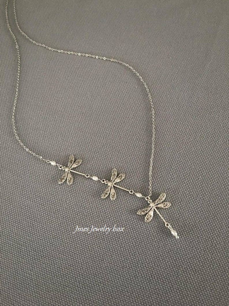 Puede incluir: Un collar de tono plateado con tres colgantes de lib&eacute;lula dispuestos en fila, acentuados con peque&ntilde;as cuentas blancas a lo largo de la cadena. El texto Jmes Jewelry box est&aacute; impreso en blanco cerca de la parte inferior del dise&ntilde;o de lib&eacute;lula.