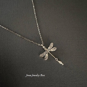May include: A silver-toned necklace featuring a dragonfly pendant with intricate filigree wing details and a small, dangling white pearl-like bead at the tail. The text Jmes Jewelry Box is printed in the lower left corner of the image.