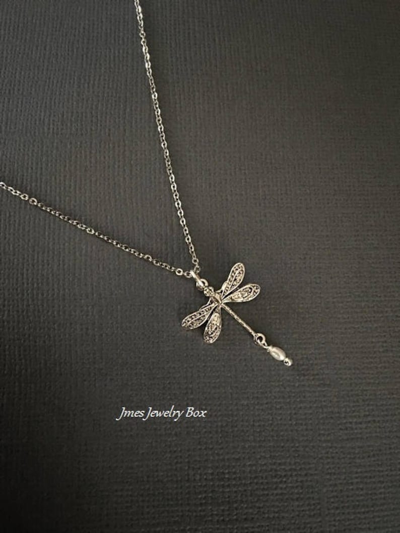 May include: A silver-toned necklace featuring a dragonfly pendant with intricate filigree wing details and a small, dangling white pearl-like bead at the tail. The text Jmes Jewelry Box is printed in the lower left corner of the image.