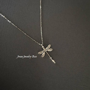May include: A silver-toned necklace featuring a dragonfly pendant with intricate wing detailing and a small dangling white bead. The text Jmes Jewelry Box is visible on the left side of the pendant. The chain is thin and metallic in appearance.