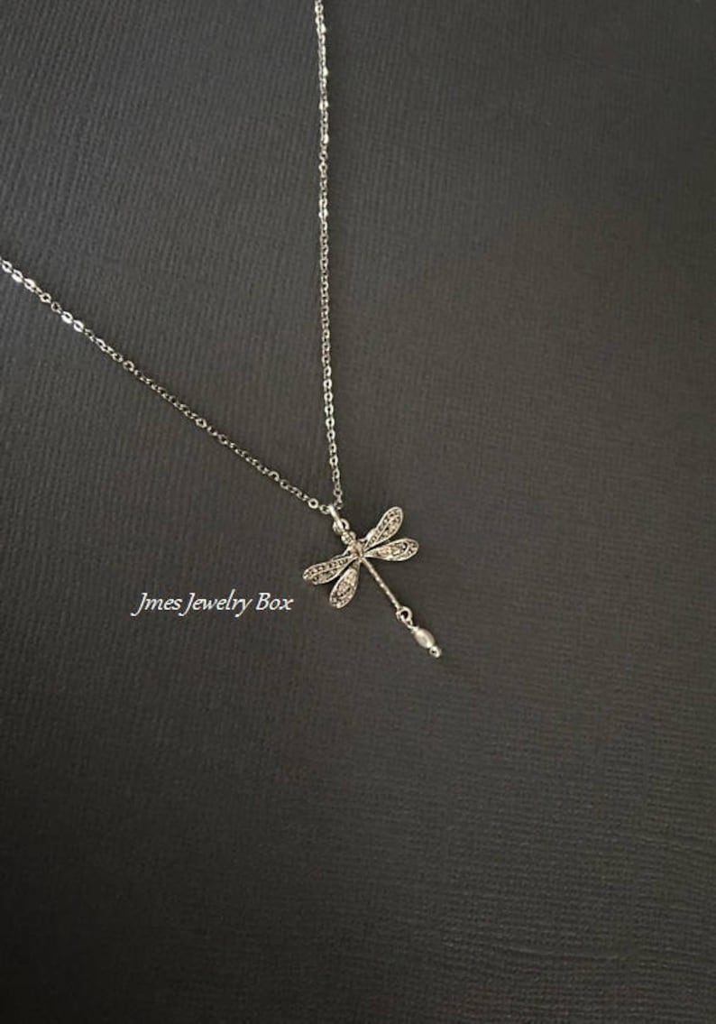 May include: A silver-toned necklace featuring a dragonfly pendant with intricate wing detailing and a small dangling white bead. The text Jmes Jewelry Box is visible on the left side of the pendant. The chain is thin and metallic in appearance.