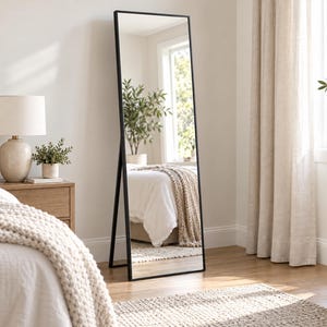May include: A tall, rectangular floor mirror featuring a thin, black metal frame. The mirror is supported by a sturdy, integrated stand that allows it to lean independently, reflecting the surrounding bedroom space and furniture.