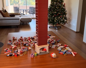 Puede incluir: Una torre de calendario de adviento alta y rectangular con patrón de ladrillo rojo, que presenta una figura de Santa Claus atrapada boca abajo en la parte superior. La base parece una chimenea y está rodeada de varios chocolates.