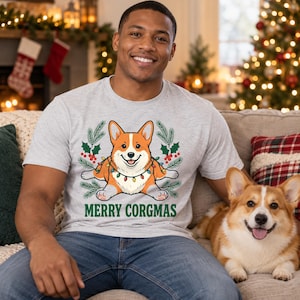 May include: A light gray short-sleeve t-shirt featuring a graphic of a Corgi dog wrapped in colorful Christmas lights, surrounded by pine branches and holly berries, with the green text MERRY CORGMAS printed below the illustration.