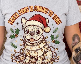 May include: A light gray t-shirt featuring a graphic of a French Bulldog wearing a red Santa hat and wrapped in a string of Christmas lights. The text above the dog reads, "SANTA PAWS IS COMING TO TOWN." Green holly leaves are on both sides.