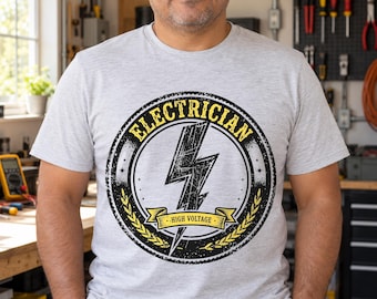 Pode incluir: Uma t-shirt cinzenta clara de manga curta com um gráfico circular no peito. O design inclui um relâmpago preto, espigas de trigo amarelas e o texto "ELECTRICIAN" e "HIGH VOLTAGE" numa fonte arrojada e desgastada.